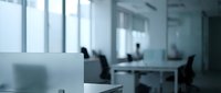Modern blurred office interior with desks, chairs and large windows creating soft natural lighting for corporate business workspace backgrounds.