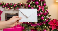 Hand holding white envelope against festive Christmas tree with pink decorations and poinsettias for holiday greeting cards and seasonal correspondence.