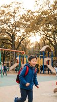 Happy Asian elementary school boy with backpack running at playground during golden hour sunset with colorful equipment and trees in background.