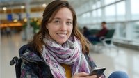 Young Caucasian woman in winter scarf smiling while using smartphone in airport terminal, waiting area with blurred passengers in background. Travel lifestyle concept.