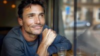 Mature Caucasian man with charming smile wearing casual sweater sitting relaxed at cafe window, hand on chin, natural daylight illuminates distinguished features.