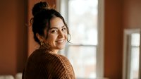 Young Hispanic woman with curly hair in cozy brown sweater smiling warmly by window in soft natural light for lifestyle and portrait photography.