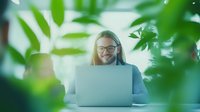 Young Caucasian woman in glasses working on laptop surrounded by green plants in bright modern office space, expressing joy and satisfaction while remote working.