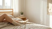 Relaxed woman legs in cozy knit socks resting on unmade bed in bright minimalist bedroom with natural morning sunlight streaming through windows.