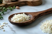 Raw white rice grains in rustic wooden spoon on marble surface with scattered rice and fresh herbs, macro food photography for cooking blogs and recipes.