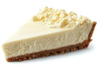 Slice of creamy vanilla cheesecake with graham cracker crust and white chocolate shavings on top, photographed against white background for food marketing.