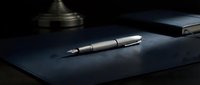 Elegant fountain pen with chrome and dark barrel resting on navy blue leather desk pad, professional writing instrument for business correspondence and executive use.