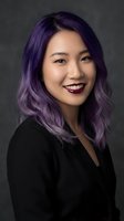 Young Asian woman with vibrant purple ombre hair and red lipstick smiling confidently in black top against dark background for professional headshot.