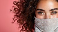 Young mixed race woman with curly hair and expressive hazel eyes wearing protective white face mask against pink background, looking directly at camera with confident gaze.