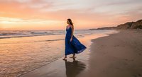 Young woman in flowing blue dress walking along ocean beach at golden sunset with waves and sandy shore creating peaceful coastal scene.