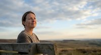 Middle-aged Caucasian woman enjoying peaceful sunset moment outdoors, leaning on wooden fence with serene expression and warm golden light on face.