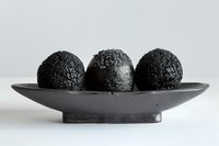 Three black charcoal spheres arranged on minimalist gray ceramic plate against white background, creating dramatic contrast and modern aesthetic composition.