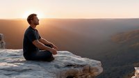 Young Caucasian man in black shirt sitting on rocky cliff edge overlooking mountain valley during golden hour sunset, peaceful meditation and outdoor adventure concept.
