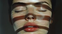 Dramatic portrait of a woman's face with closed eyes and red lips illuminated by striped light and shadow pattern creating artistic geometric effect on skin.