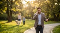 Professional businessman talking on smartphone while walking in sunny park with family in background during golden hour.