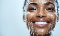 Young African American woman with bright smile splashing water on face during skincare routine, showcasing fresh, glowing, hydrated skin in closeup view.