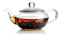 Glass teapot with brewing loose black tea leaves floating in amber liquid, transparent design shows steeping process against white background. Isolated.