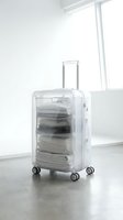 Transparent hard-shell suitcase packed with neatly folded white clothes, standing in a bright minimalist room with concrete floor.