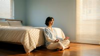 Woman with dark hair and medium skin tone sitting cross-legged on wooden floor in bedroom wearing light cardigan, meditating or contemplating by bed with white bedding and window blinds, suitable for wellness and lifestyle content.