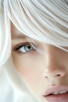 Close-up portrait of young woman with platinum blonde hair partially covering intense blue eye, natural makeup and soft pink lips against light background.