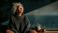 Mature African American woman with curly gray hair in cozy sweater enjoying peaceful moment with pottery and warm lighting for lifestyle content.