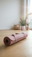 Pink yoga mat rolled up with leather straps on wooden floor in bright minimalist room with plants and natural sunlight for fitness and wellness.