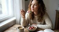 Young Caucasian woman enjoying healthy breakfast bowl with berries and granola at home kitchen table by window, morning wellness lifestyle concept.