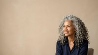 Mature woman with long gray curly hair and mixed ethnicity wearing dark blazer, smiling while looking away against neutral beige background for corporate and lifestyle use.