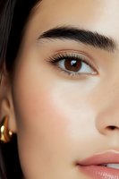 Close-up of Asian woman's face showing natural makeup, groomed eyebrow, brown eye with long lashes, and glossy nude lips, wearing gold earring.