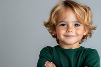 Confident young child with blonde wavy hair wearing green sweater, arms crossed, showing genuine smile against light gray background. For family, education, childhood concepts.