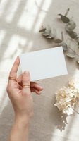 Female hand holding blank white card mockup with eucalyptus and dried flowers on neutral background with soft shadows for branding design.