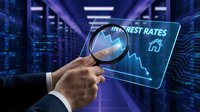 Hand holding magnifying glass over transparent digital interface showing interest rates chart with declining arrow in modern server room background.