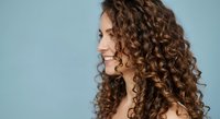 Young woman with voluminous curly brown hair in profile view against soft blue background, natural beauty and haircare concept for salon advertising.