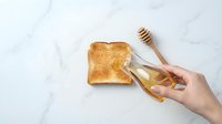 Caucasian hand holding wooden honey dipper over golden toast on marble surface, breakfast preparation and natural sweetener application.
