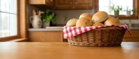 Fresh baked bread rolls in wicker basket with checkered cloth on wooden kitchen counter. Warm homemade bakery goods for breakfast or dinner preparation.
