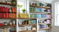 Organized home library with colorful books arranged by color on wooden shelves creating rainbow effect in bright modern living room interior design.