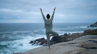 Person in warrior pose on rocky coastal cliff overlooking ocean waves, practicing yoga or fitness routine outdoors in natural seaside setting.