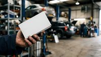 Male mechanic holding blank white business card in auto repair shop garage with vehicles and equipment in background.