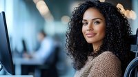 Young African American businesswoman with curly hair smiling at workplace, wearing casual sweater. Professional female employee at modern office desk with computer.