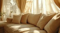 Cozy beige sofa with soft cushions in sunlit living room, morning light streaming through sheer curtains creating warm atmosphere and shadow patterns.