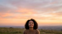 Woman with curly dark hair wearing tan sweater gazes upward at golden sunset sky over rolling landscape hills, conveying peace and contemplation.