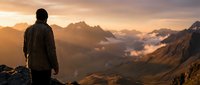 Male hiker silhouette standing on mountain peak during golden hour sunrise, contemplating dramatic alpine landscape with misty valleys below.