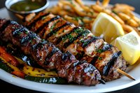 Grilled beef kebab skewer with charred marks, served with french fries, fresh lemon wedges, and herbs on white plate, accompanied by dipping sauce.