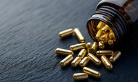 Golden dietary supplement capsules spilling from brown glass bottle onto dark slate surface, dramatic side lighting creates contrast and shadows. For health content.