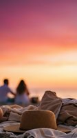 Straw hat rests on sandy beach with couple sitting by ocean during golden sunset, perfect for travel and lifestyle marketing.
