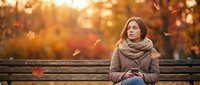 Young woman in cozy scarf sitting on wooden bench during golden autumn sunset with falling leaves creating warm atmospheric bokeh background.
