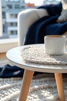 Coffee cup on knitted coaster atop modern scandinavian side table with navy throw blanket and textured rug creating cozy home atmosphere.