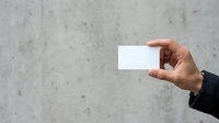 Male hand with medium skin tone holding blank white business card against concrete wall background for corporate branding mockup.