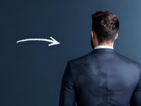 Businessman in dark suit viewed from behind looking at white arrow drawn on navy blue background, representing direction, guidance and future opportunities.