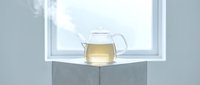 Glass teapot filled with hot herbal tea on concrete cube against bright window, minimalist beverage setup for wellness and relaxation themes.